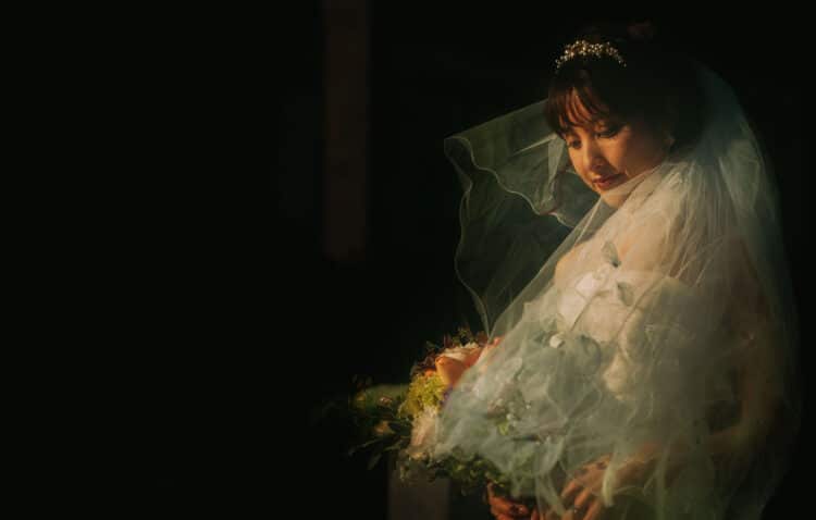 Takuya, a bride wearing a veil and holding a bouquet, stands elegantly.