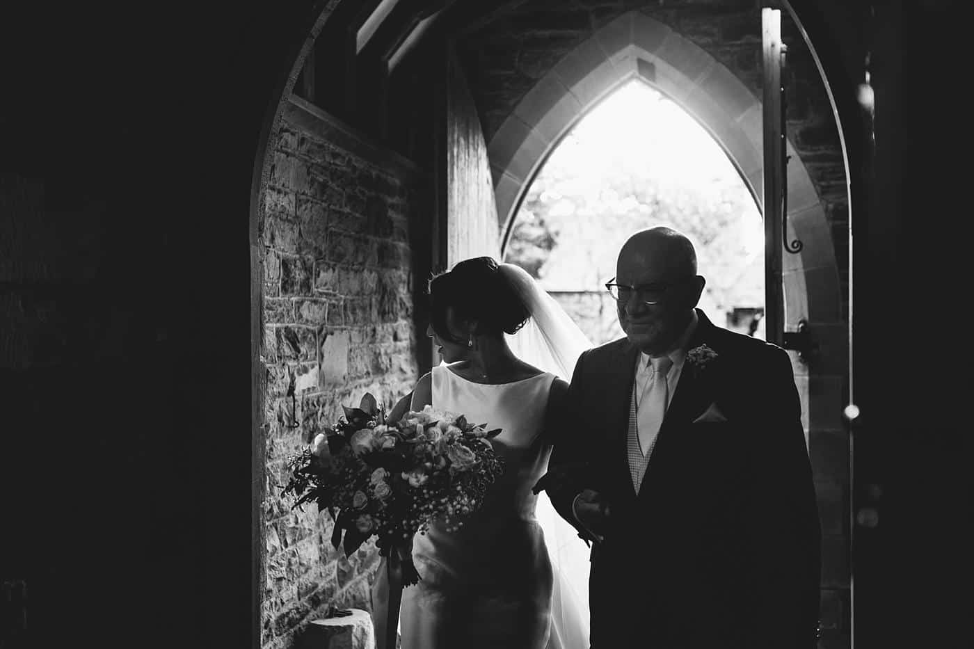 A black and white photo of a bride and her father walking through an archway during their Wistanstow Village Hall wedding.