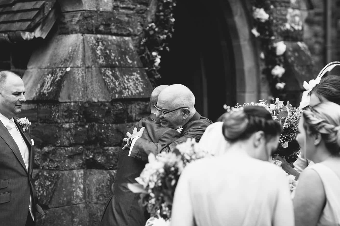 A bride and groom hugging in front of Wistanstow Village Hall.