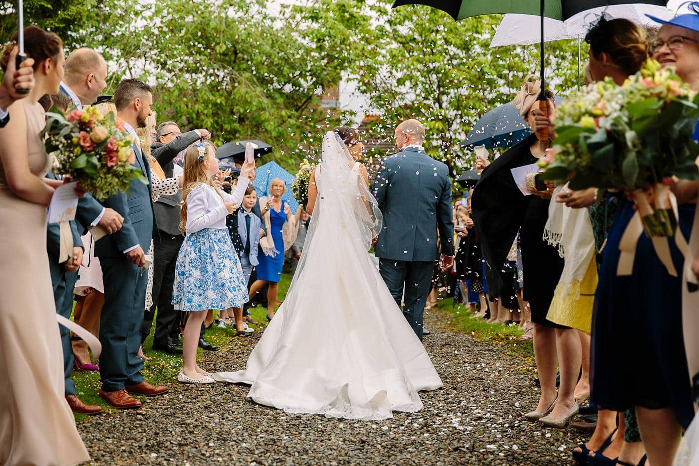 A bride and groom walking down a path with umbrellas during their Wistanstow Village Hall Wedding.