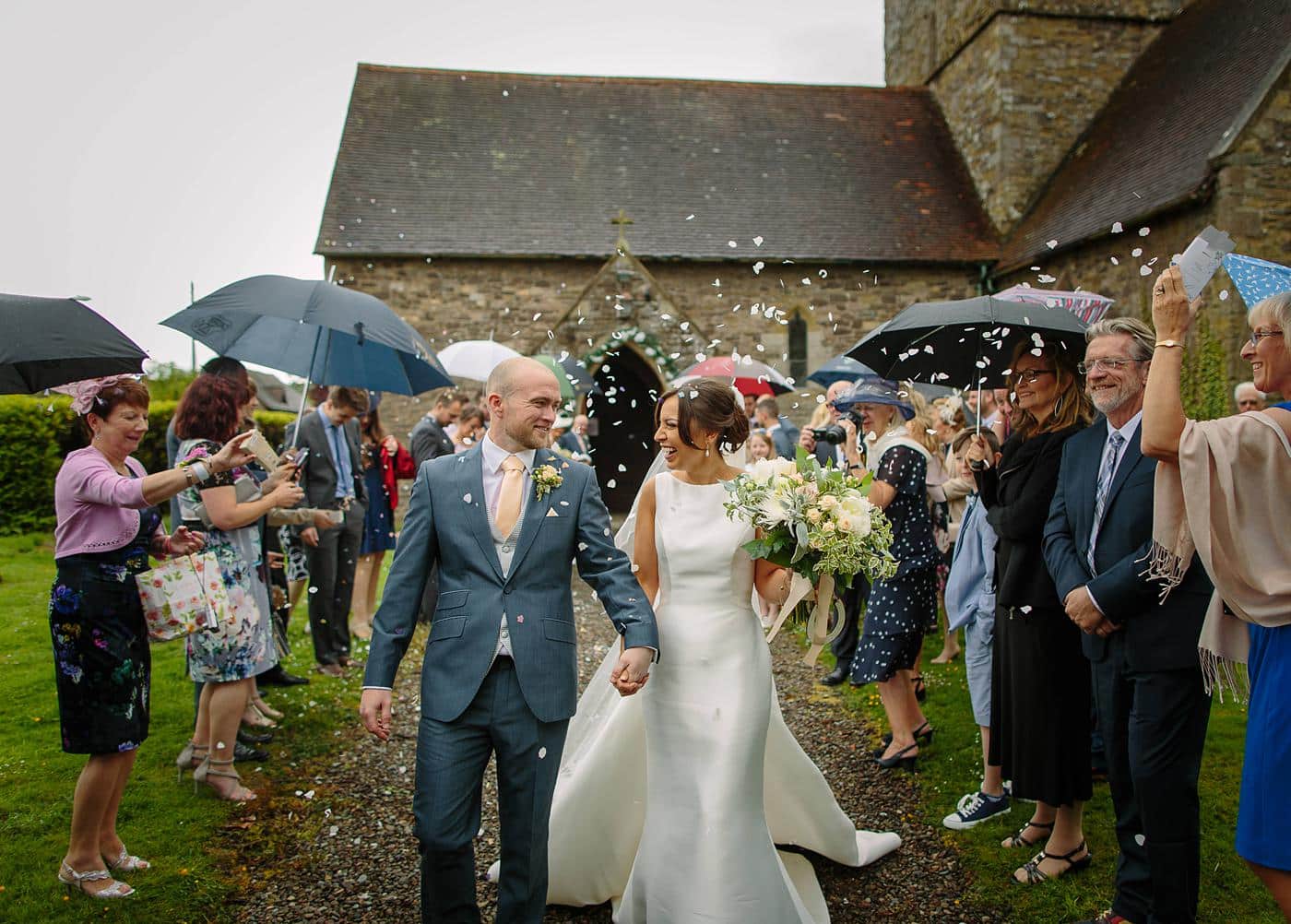 A bride and groom walk down a path at their Wistanstow Village Hall wedding, with confetti thrown at them.