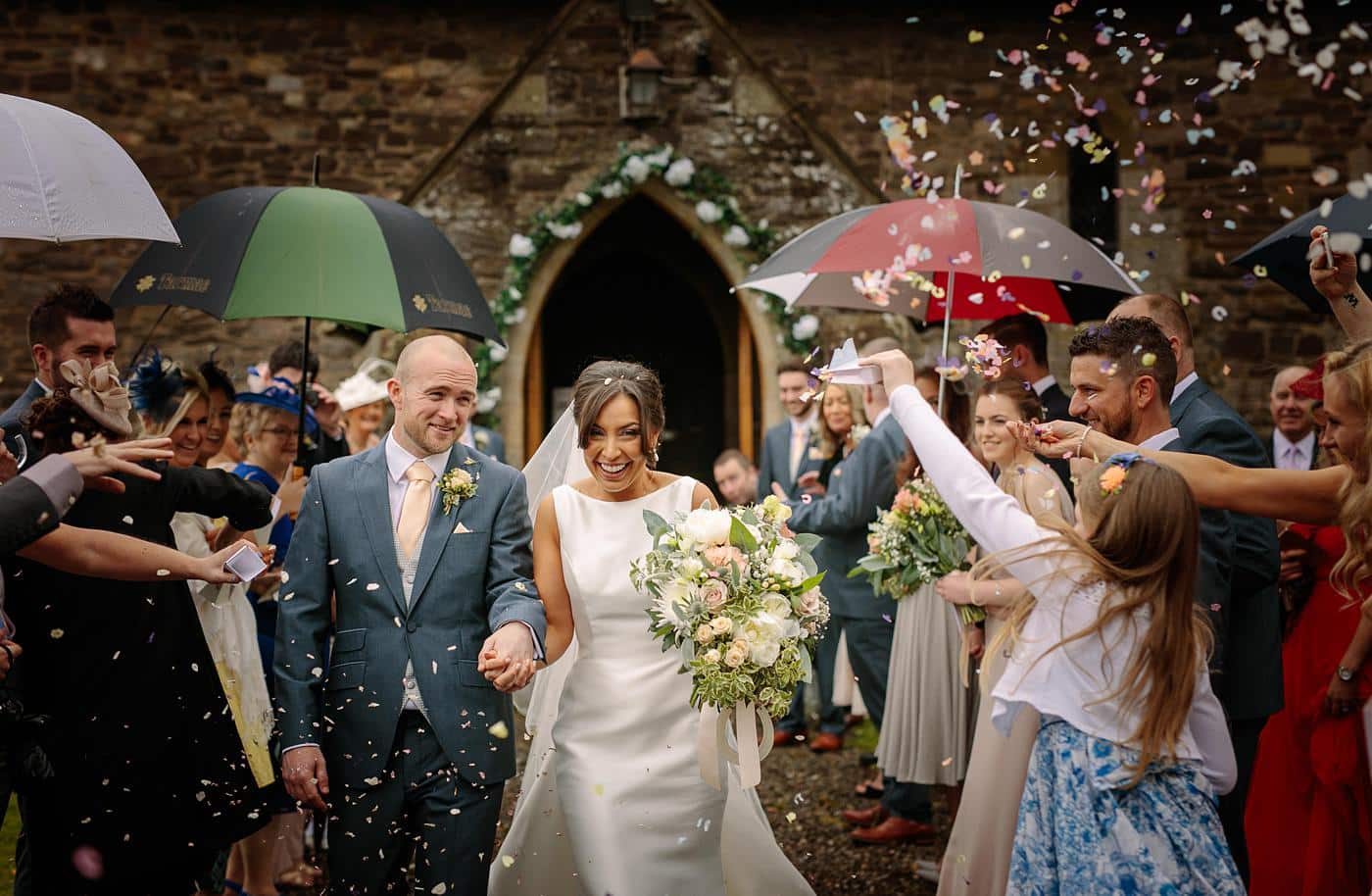 A bride and groom walk down the aisle at their enchanting Wistanstow Village Hall wedding, surrounded by a joyful cascade of confetti.