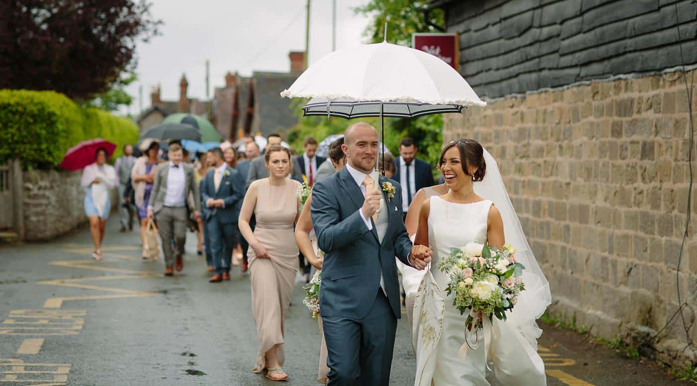 A bride and groom walking down a street in Wistanstow Village Hall with umbrellas.