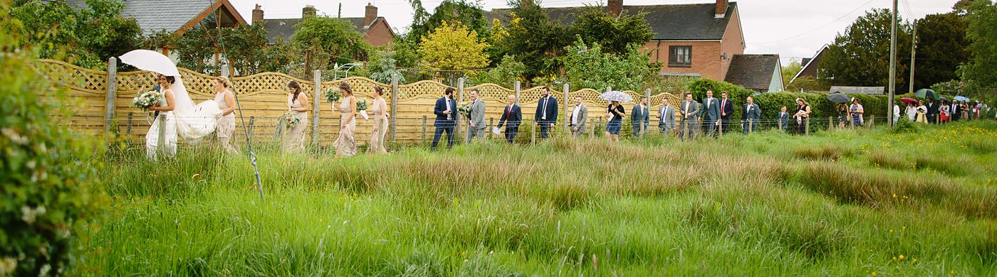 A group of people standing in a Wistanstow Village Hall, surrounded by a grassy field at a wedding celebration.