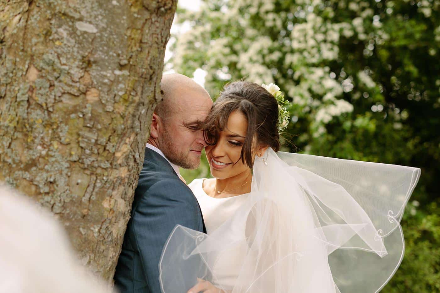 A bride and groom hugging in front of a tree at their Wistanstow Village Hall wedding.
