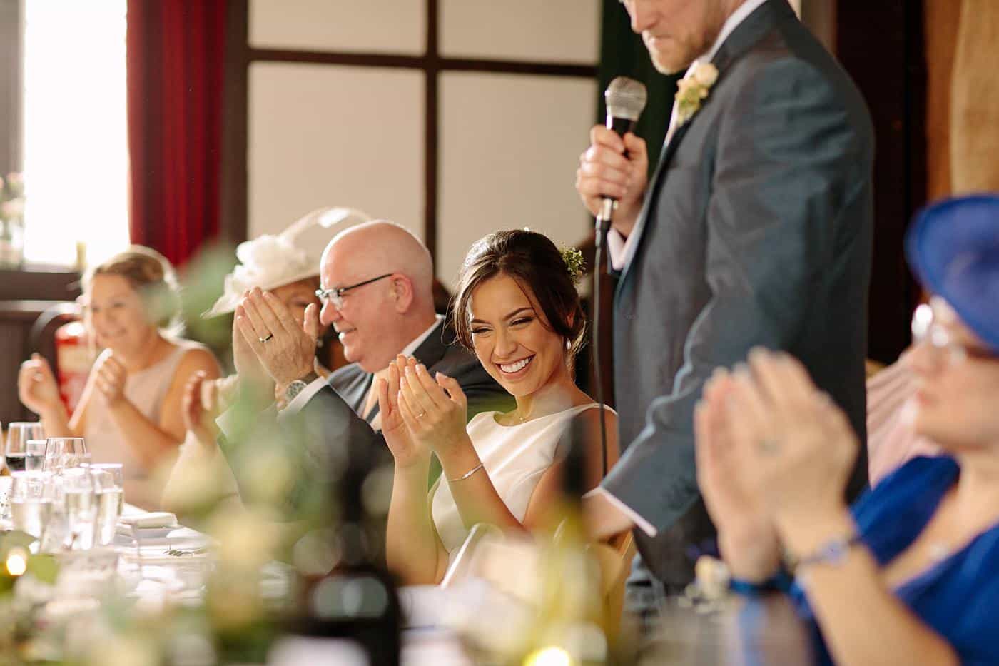 The newlyweds, surrounded by their loved ones, joyfully clapping at their wedding reception held at Wistanstow Village Hall.