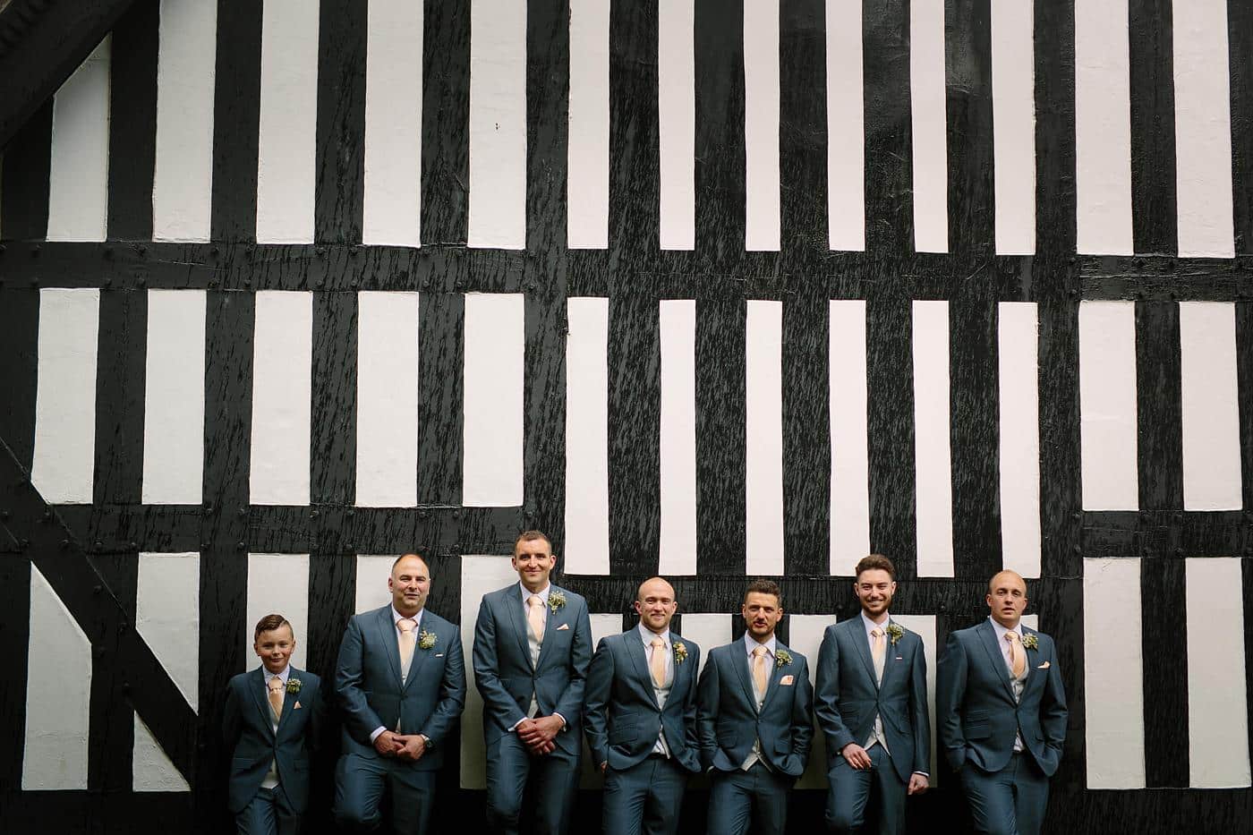 A group of groomsmen standing in front of Wistanstow Village Hall at a wedding.