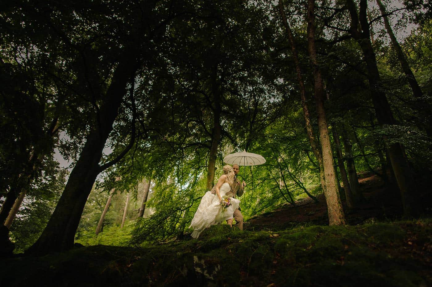 On their wedding day, a bride and groom stand beneath an umbrella, surrounded by the enchanting beauty of the woods.