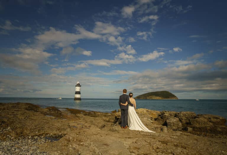 A bride and groom standing on rocks near a lighthouse at the Tros Yr Afon Wedding venue, captured by a talented North Wales Wedding Photographer.