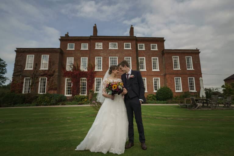 A bride and groom standing in front of a large red brick house.