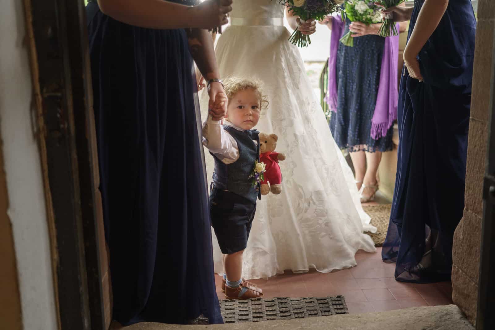 a little boy holding a teddy bear in front of bridesmaids.