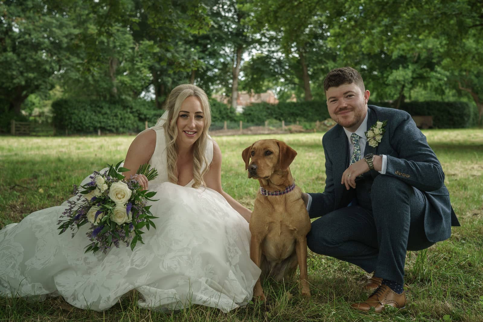 a bride and groom pose with their dog in a field.