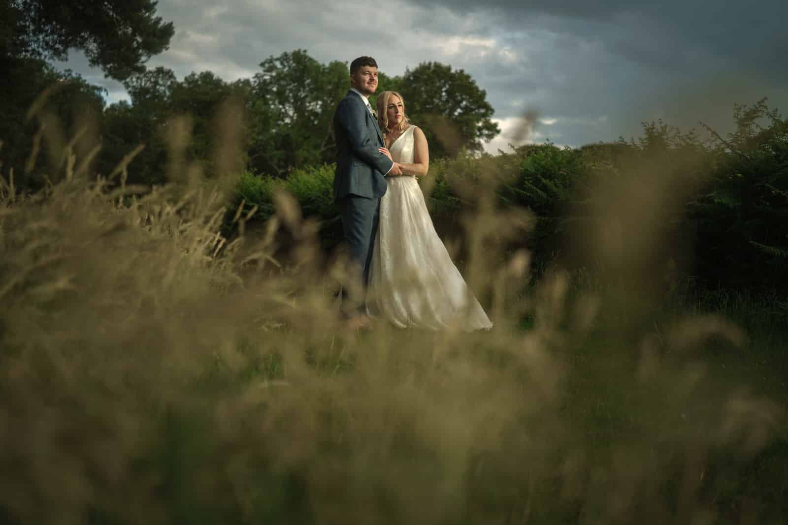 a bride and groom standing in tall grass under a stormy sky.