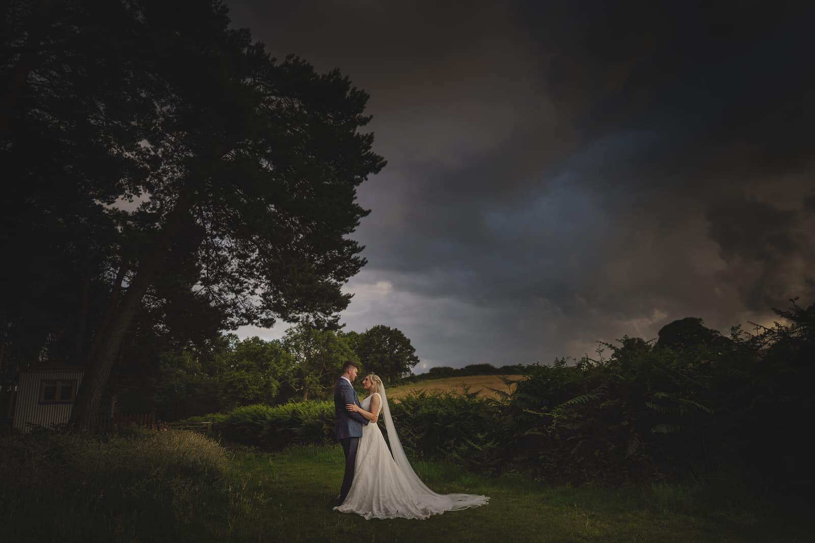 a bride and groom standing in a field under a stormy sky.