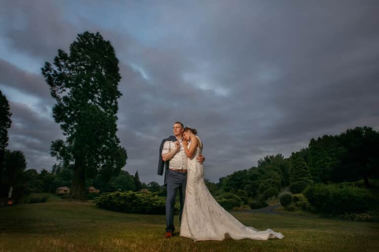a bride and groom embracing in a field near foxtail barns under a cloudy sky.