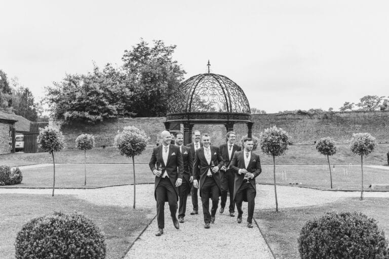 a black and white photo of a group of groomsmen walking through a garden at foxtail barns.