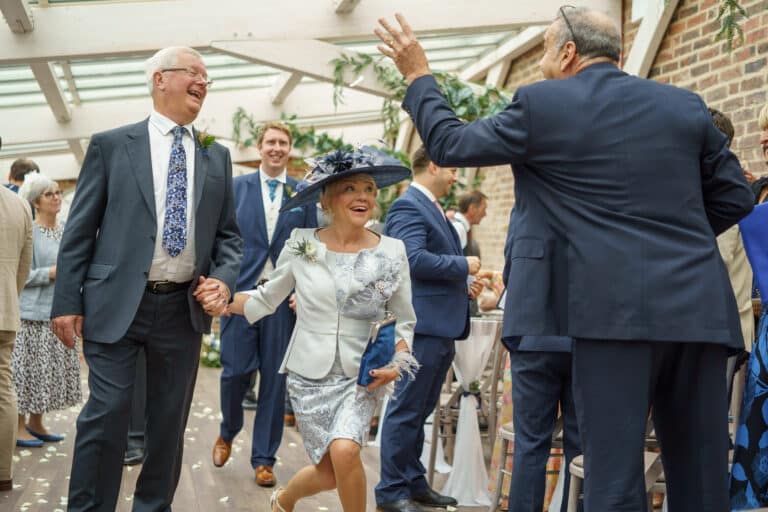 a woman in a suit and hat walks down the aisle at a wedding, set against the rustic charm of a foxtail barn.