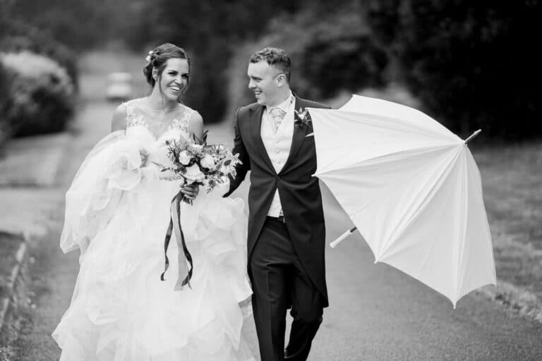 a bride and groom walking down a path at foxtail barns, gracefully holding an umbrella to shield themselves from the rain.