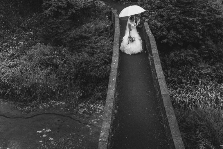 a bride and groom standing on a bridge at foxtail barns, sheltered under an umbrella.