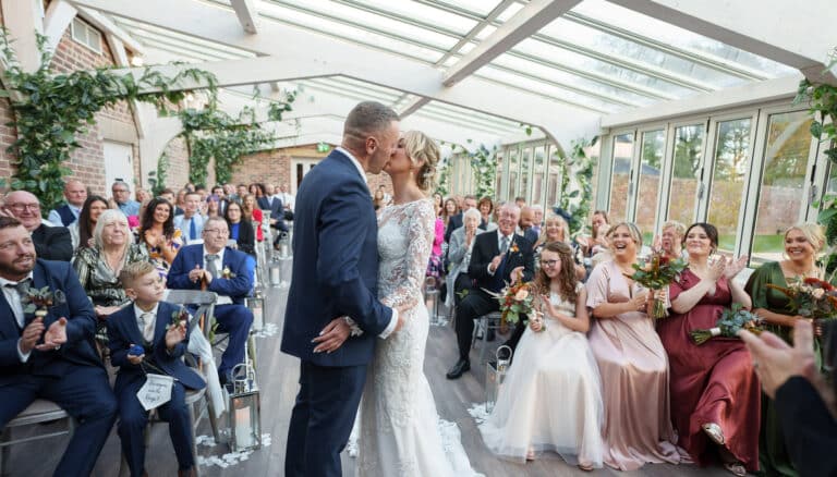 a bride and groom kiss during their wedding ceremony in the orangery at Foxtail barns, surrounded by their family and friends