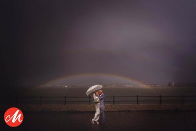 a bride and groom standing under an umbrella on their wedding day in front of a rainbow.
