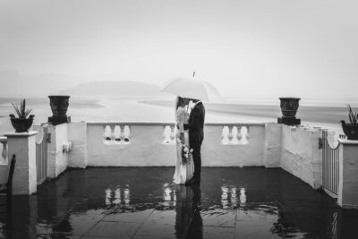 a bride and groom embracing on a rainy wedding day, sheltered by an umbrella.
