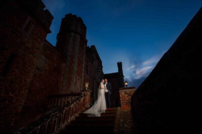 pbartworks photography captures a magical moment of a bride and groom on the steps of a castle at dusk, showcasing their skills as an exceptional wedding photographer in shropshire.