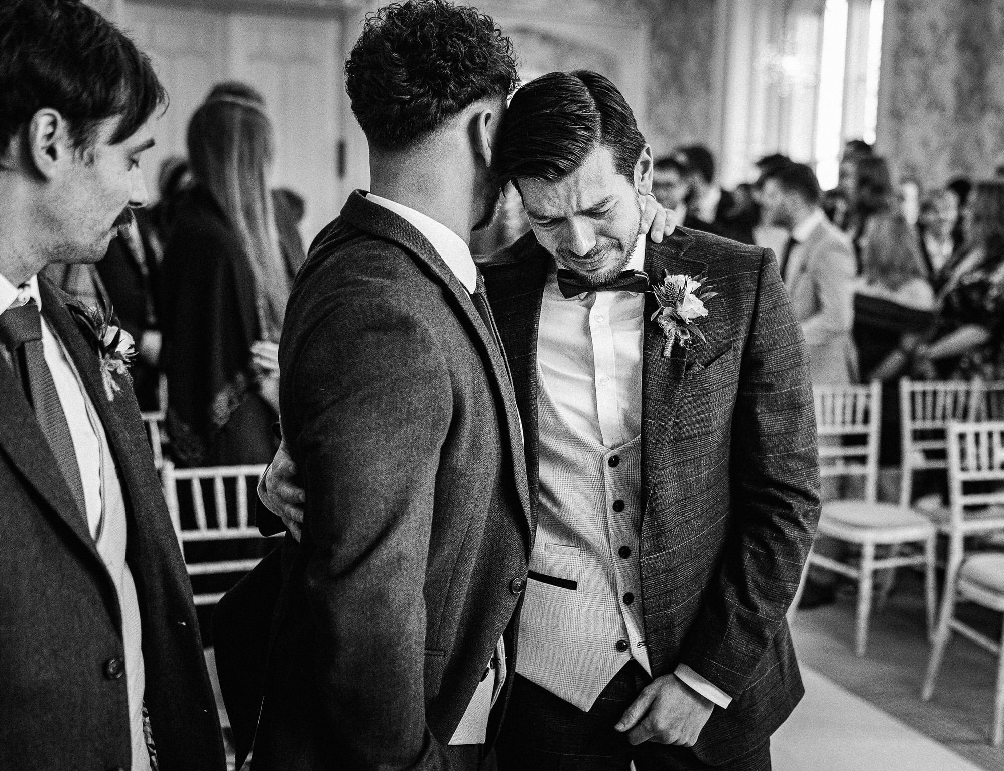 in a room filled with seated and standing people, a man consoles another man who appears emotional and is dressed in a three piece suit with a bow tie. it's a tender moment captured beautifully by pbartworks photography, the trusted wedding photographer in shropshire.