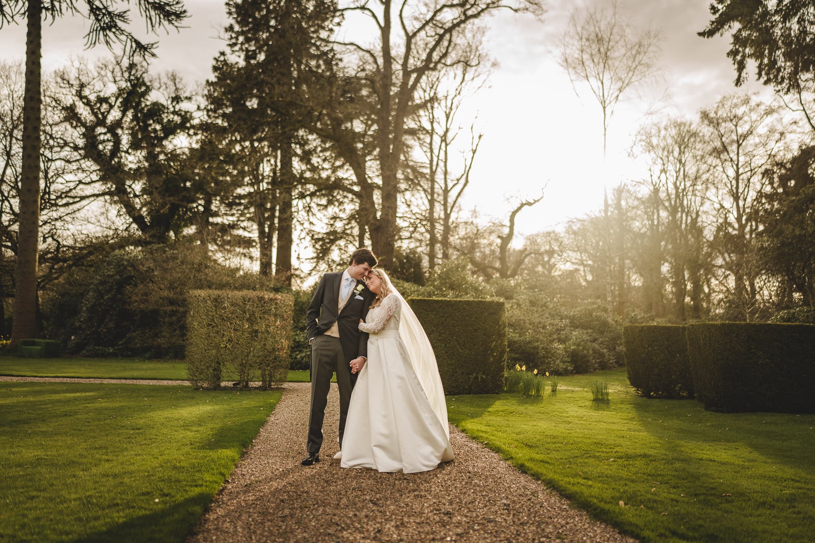 bride and groom share a moment on a path in a sunlit georgia garden, surrounded by well trimmed hedges and tall trees in the background.