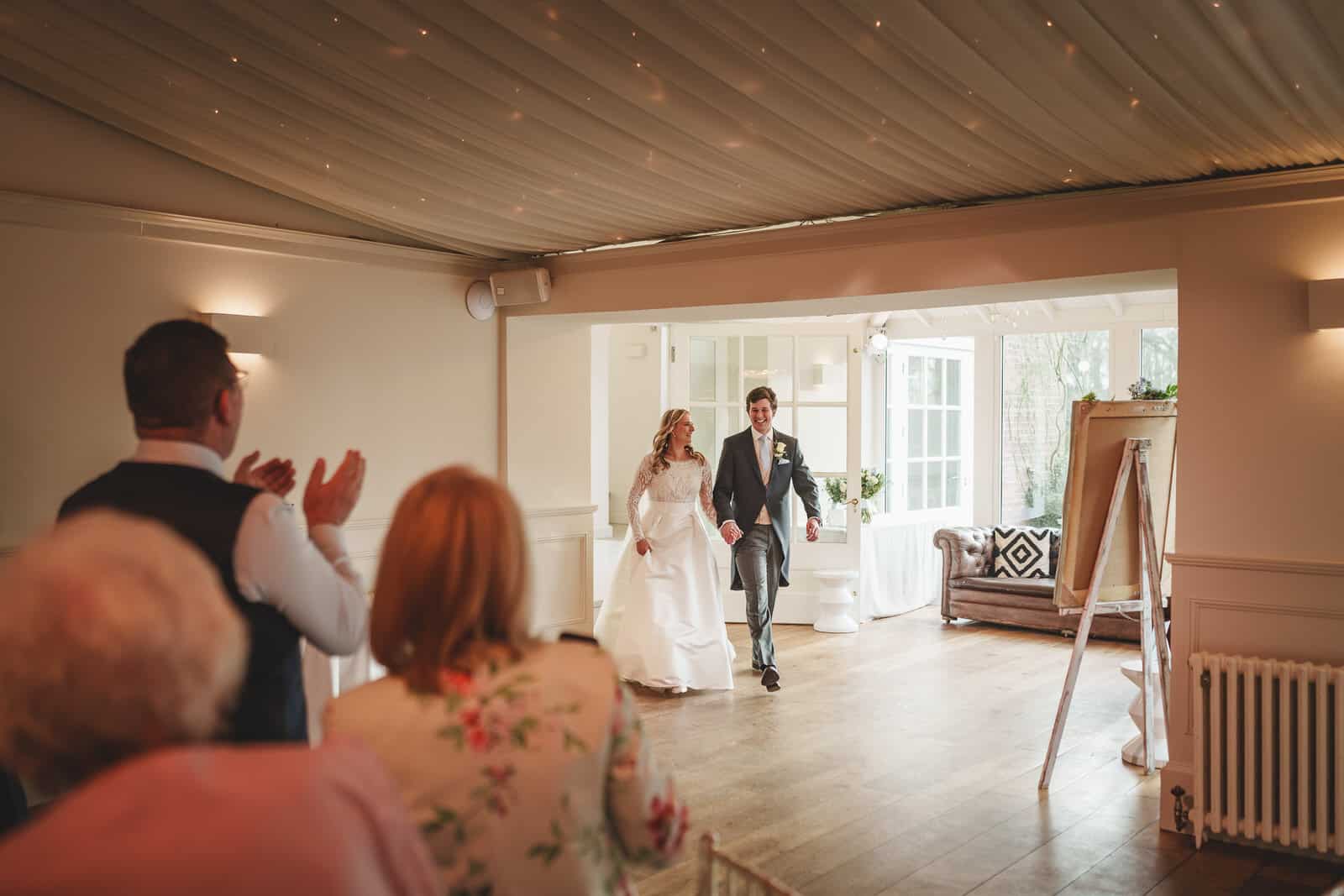 a bride and groom walk hand in hand into a well lit room, the charming wedding scene set in georgia, as guests seated on the left side applaud their entrance.