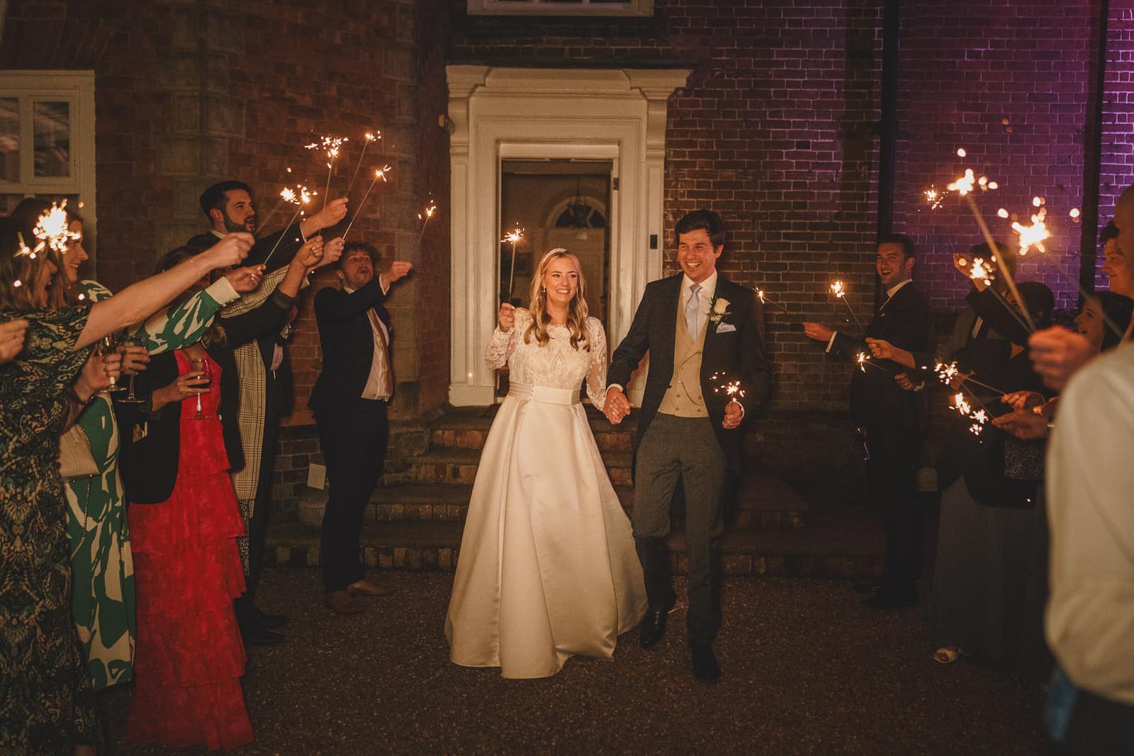 a bride and groom walk outside holding hands, smiling, surrounded by guests holding sparklers at night. the bride wears a white gown, and the groom is in a suit. this charming wedding in georgia radiates joy and elegance.