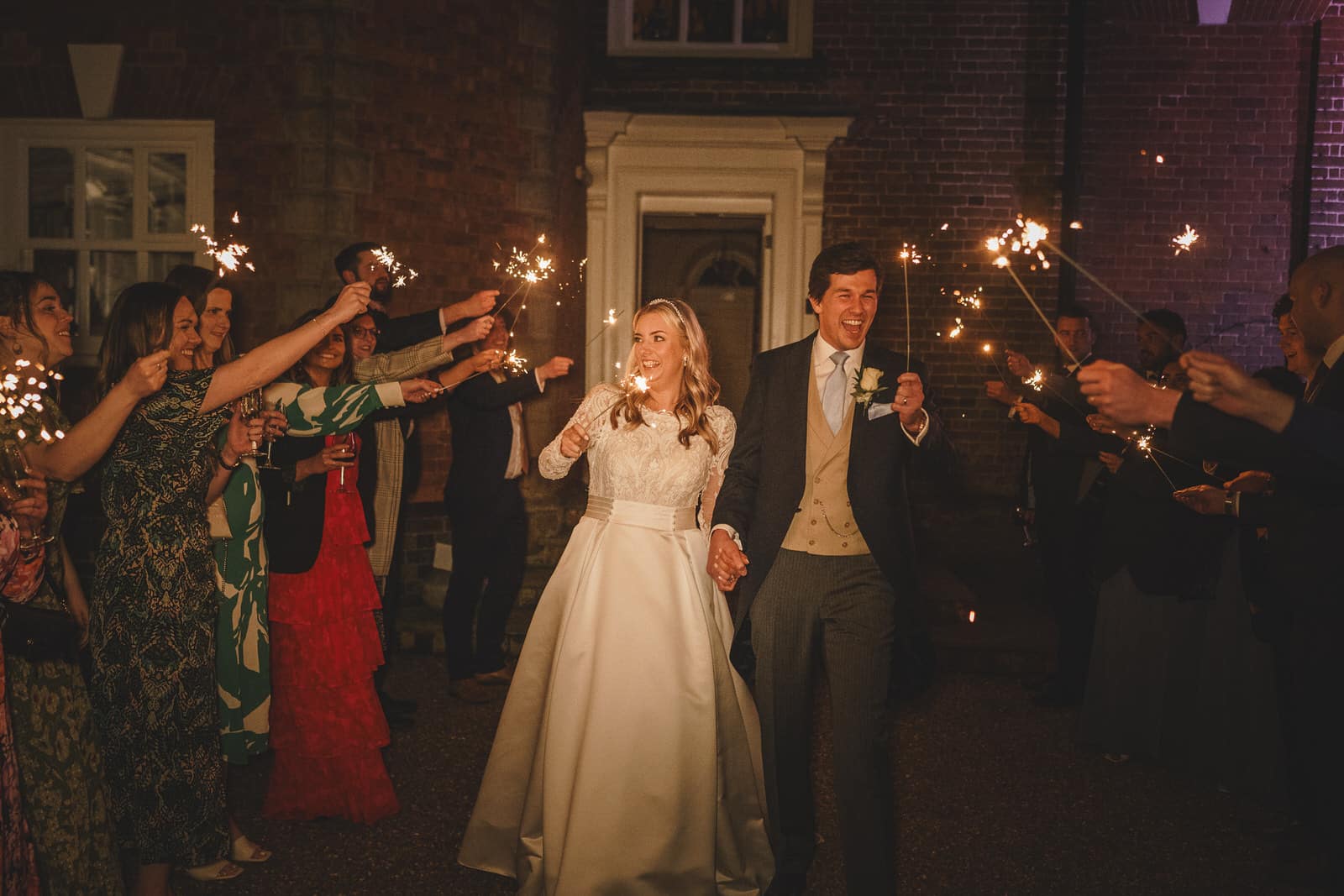 hamish and georgia walk hand in hand at night, surrounded by wedding guests holding sparklers outside a building.