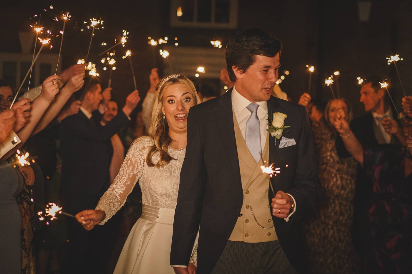 a bride and groom are walking together, smiling, and holding sparklers at their enchanting evening wedding. they are surrounded by people also holding sparklers, celebrating under the night sky.