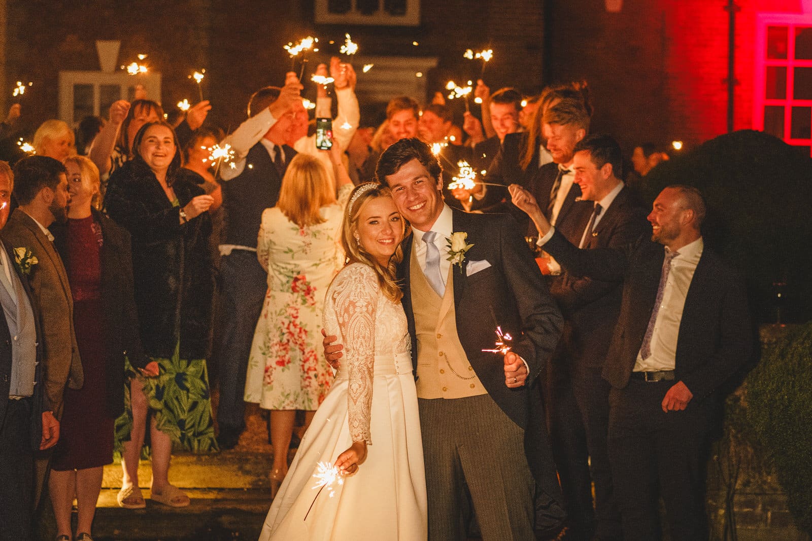 a couple, dressed in their wedding attire, stands smiling and holding sparklers amidst a crowd of people during a charming wedding celebration under the georgia night sky.