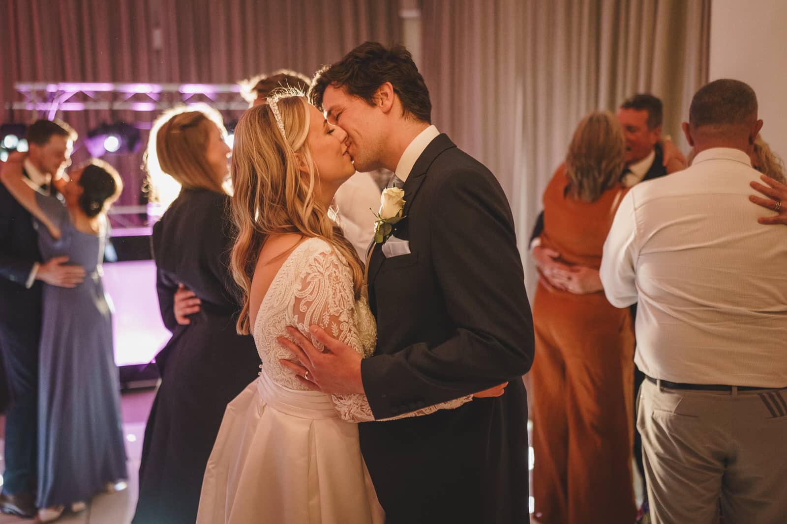 a bride and groom share a kiss on the dance floor at their georgia wedding, surrounded by couples dancing. the bride is wearing a white lace dress and the groom, hamish, is in a dark suit. the background is lit with purple lights.