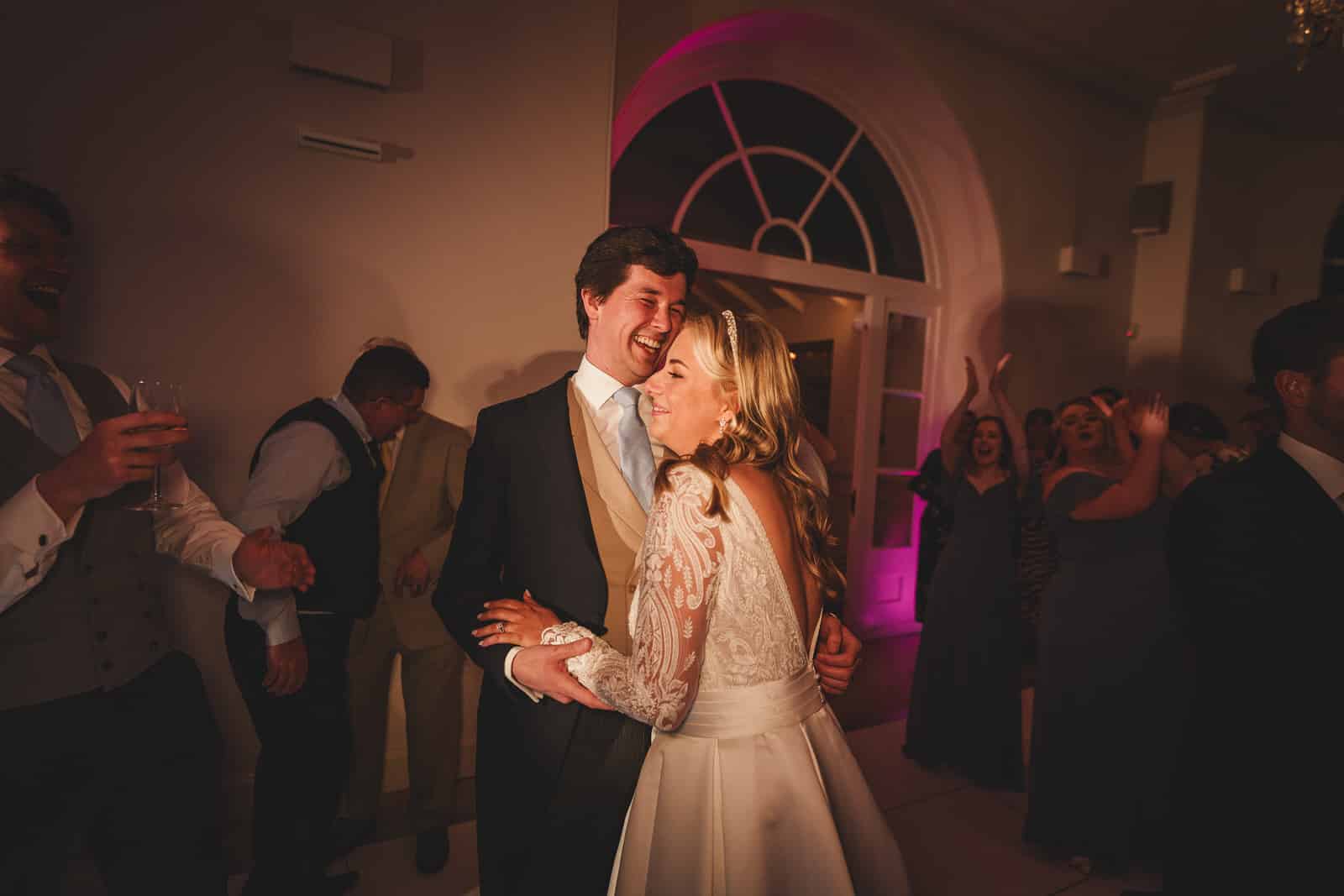 hamish and georgia share a joyful embrace at their charming wedding reception, surrounded by cheering guests in a dimly lit room with a pink light accent.