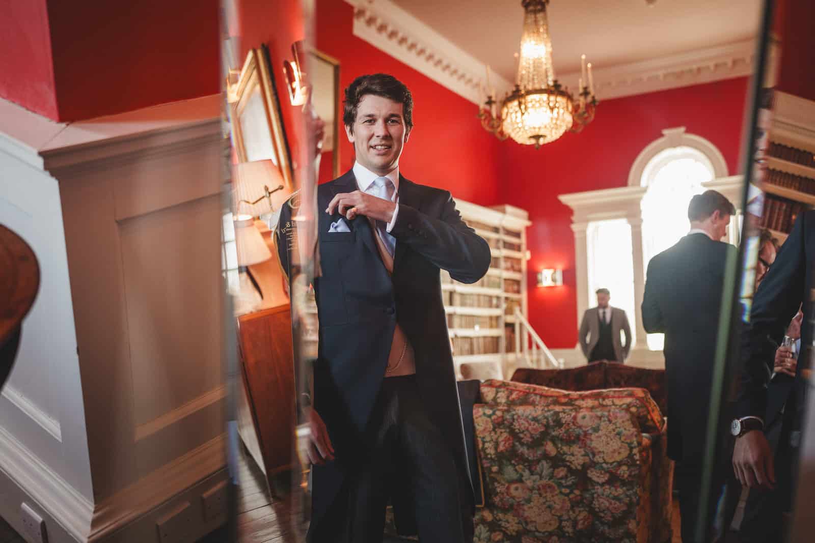 hamish, dressed in a sharp suit, adjusts his pocket square in front of a mirror in an elegant room with red walls and chandeliers, preparing for the wedding.