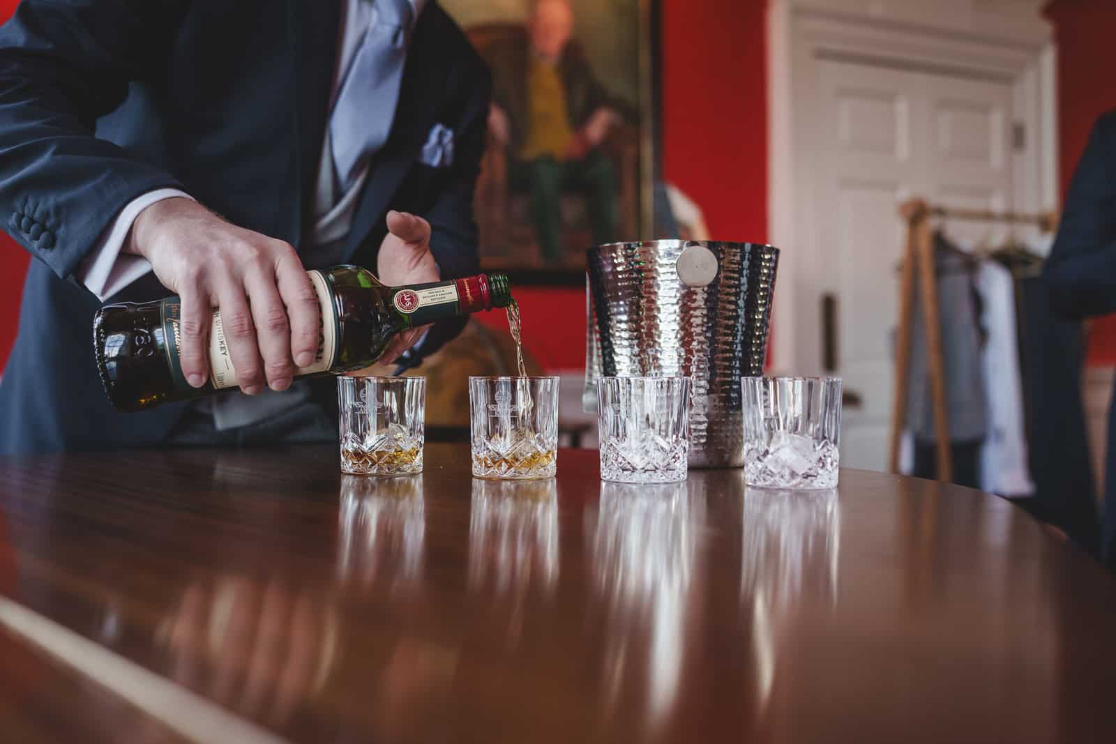 Hamish, dressed sharply in a suit, pours whiskey from a bottle into one of four glasses, three with ice, on a wooden table. In the background, a silver ice bucket and coat rack add to the setting's charm, making it perfect for Georgia’s charming wedding preparations.