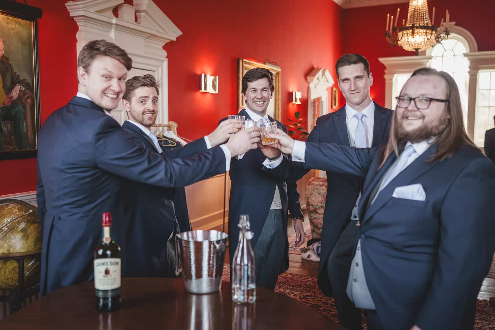 Five men in suits raise their glasses for a toast in a stylish room with red walls, celebrating Hamish's charming wedding. A bottle of whiskey, an ice bucket, and a shaker are on a table in the foreground.