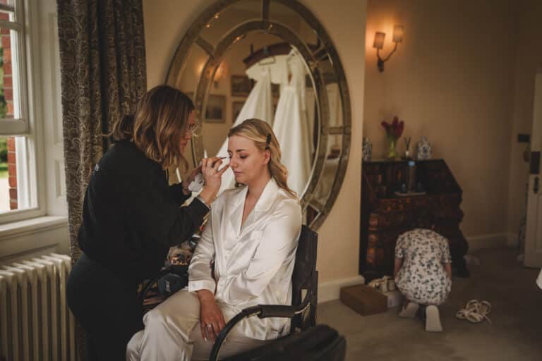A woman in a white robe sits on a chair getting her makeup done by another woman at Iscoyd Park. A wedding dress hangs in the background, and a person is kneeling near the furniture, adding to the serene atmosphere of this Shropshire setting.