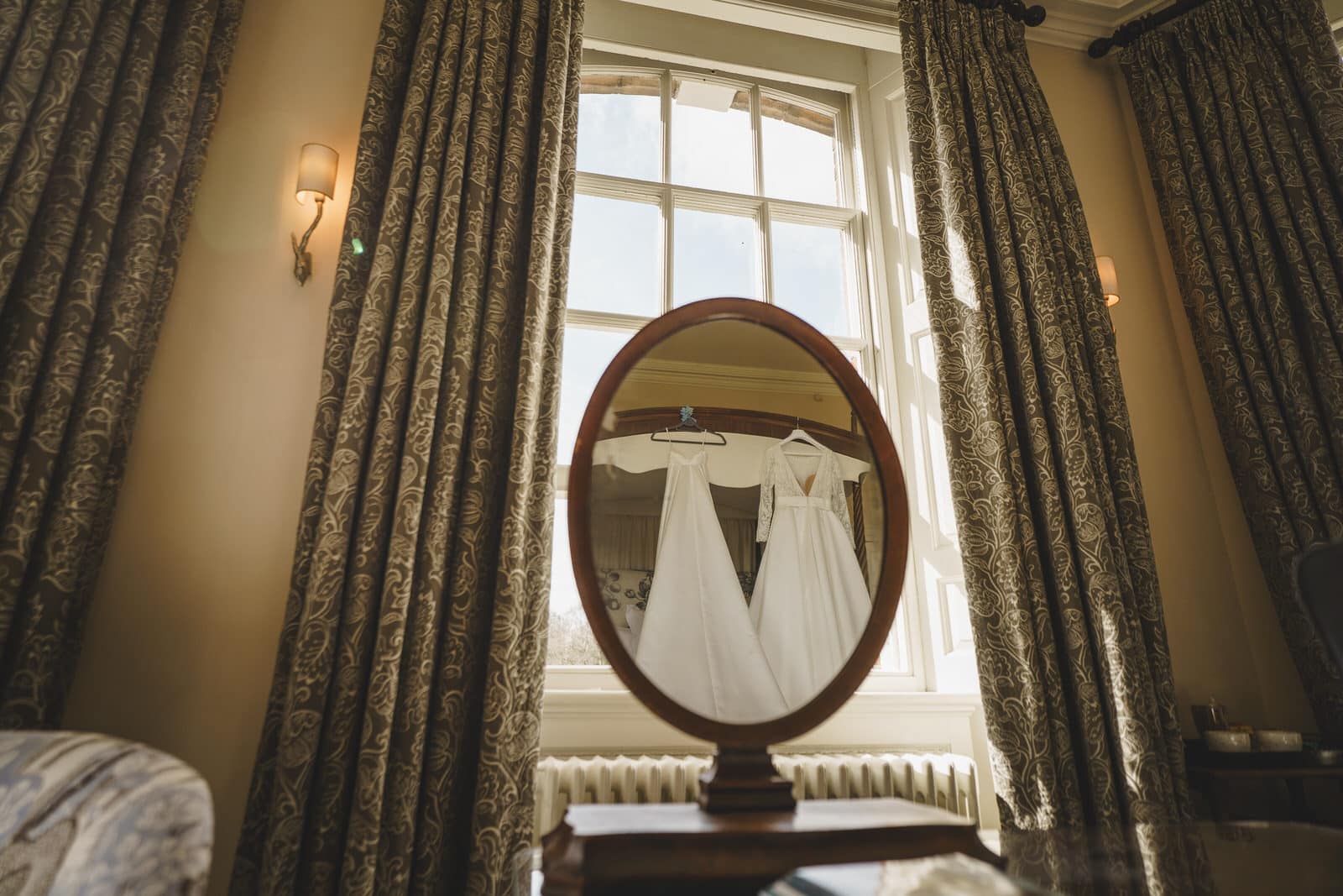 A mirror reflects two white wedding dresses hanging in front of a large window, framed by patterned curtains in a warmly lit room at Iscoyd Park, Shropshire.