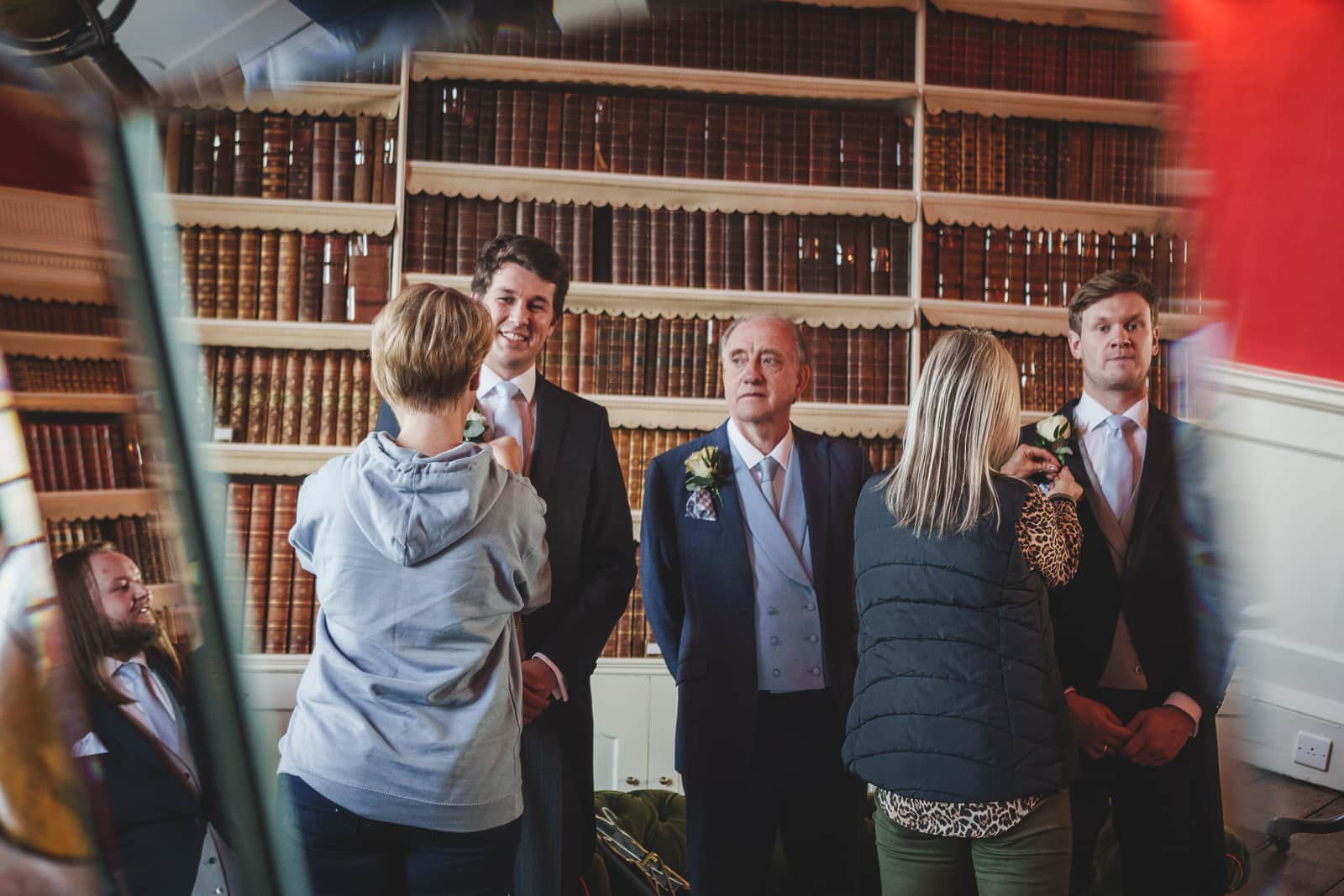 A group of people in formal attire stands in a library with wooden shelves filled with books. Georgia and Hamish are adjusting the men's outfits, preparing for the wedding ceremony.
