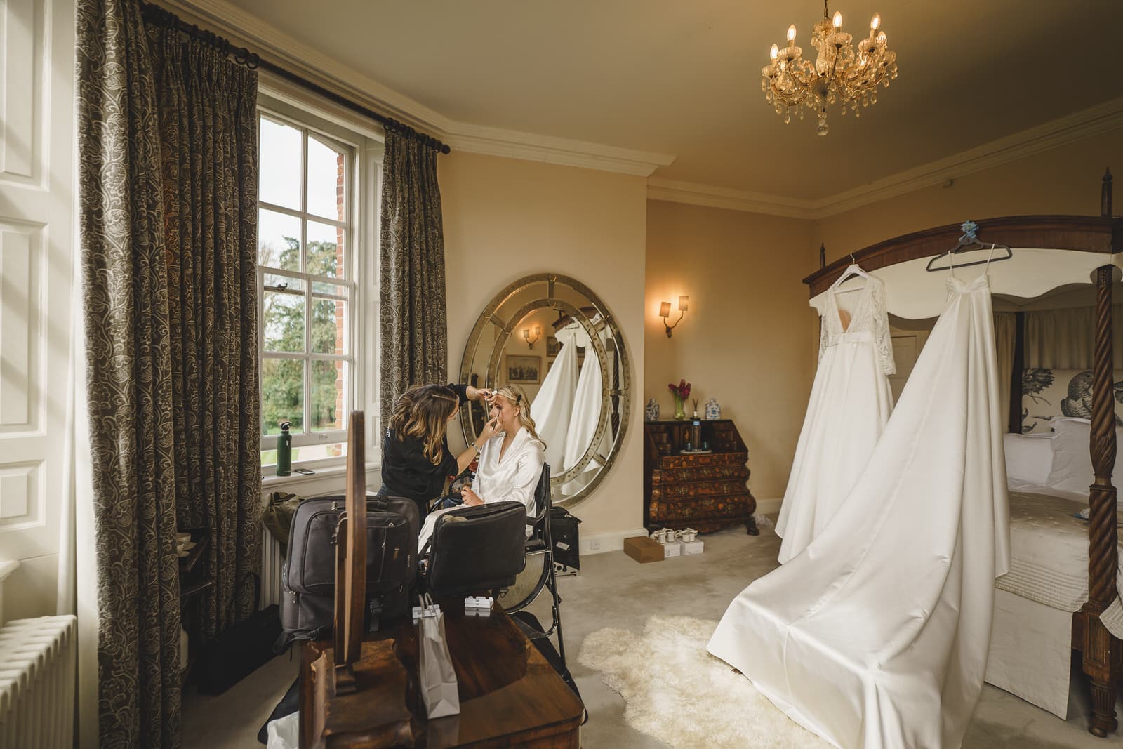 A makeup artist delicately applies makeup to a woman seated by a window in a well-decorated bedroom at Iscoyd Park, with her wedding dress hanging on the canopy bed.