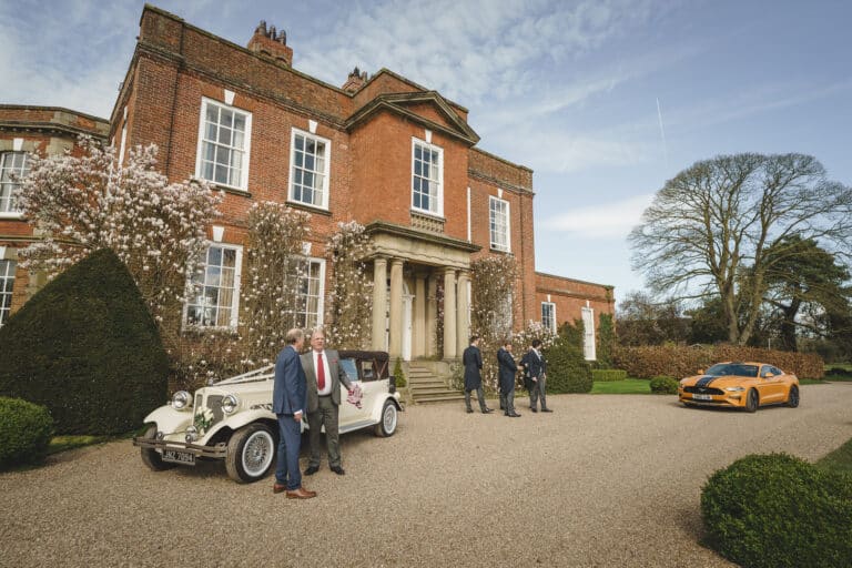 A group of people stands outside the grand brick mansion at Iscoyd Park, with a white classic car and an orange sports car parked in front. The scene is set on a clear day with blue Shropshire skies.