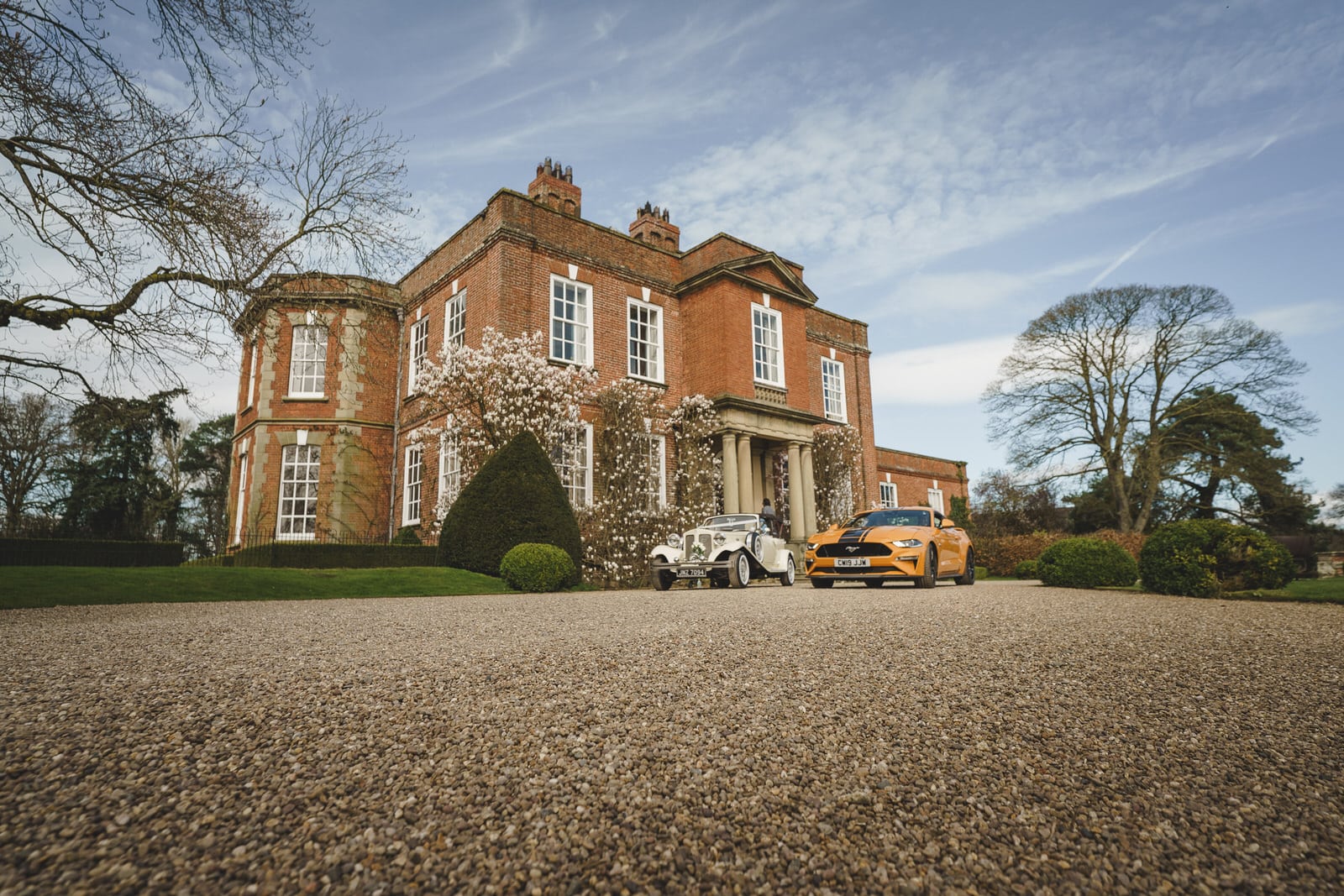 A large brick mansion at Iscoyd Park in Shropshire features two cars, one white and one yellow, parked on a gravel driveway. The mansion is surrounded by a well-kept lawn and various trees.
