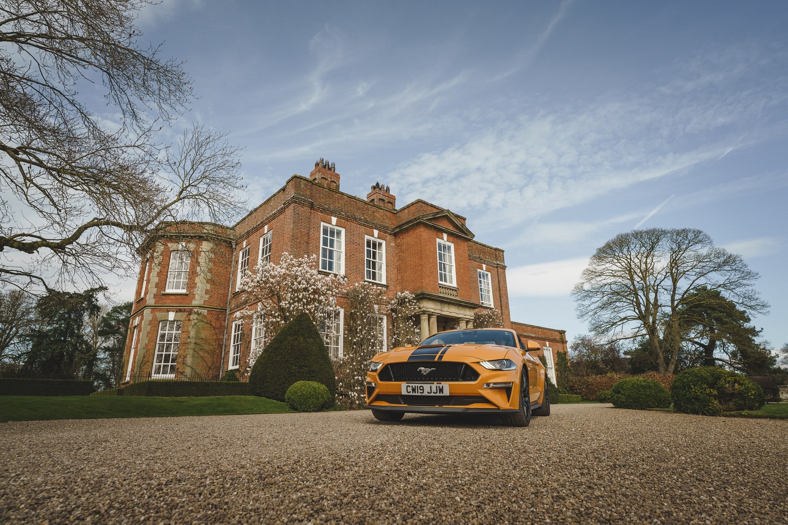 An orange Ford Mustang is parked on a gravel driveway in front of a large, red-brick mansion at Iscoyd Park, Shropshire, with a well-manicured garden and clear blue sky.