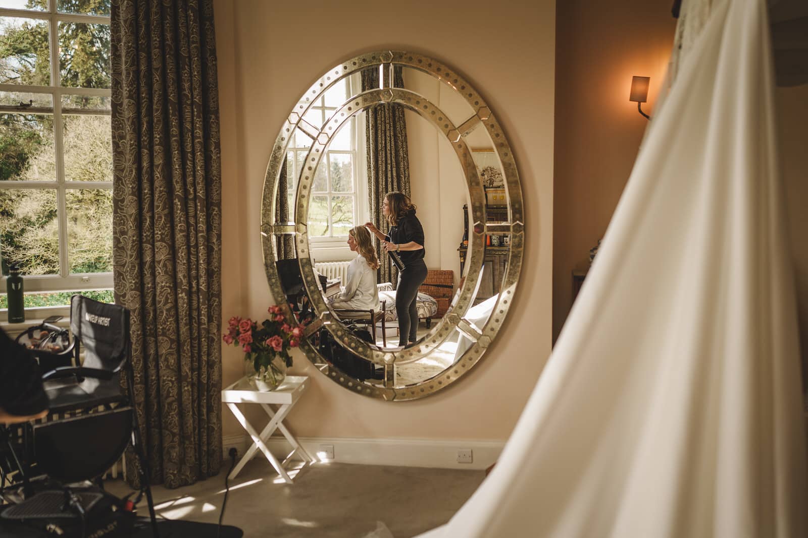 A stylist is seen through a large oval mirror working on a woman's hair in a well-lit room with tall curtains and a window at the charming Iscoyd Park in Shropshire. A dress is visible in the foreground.