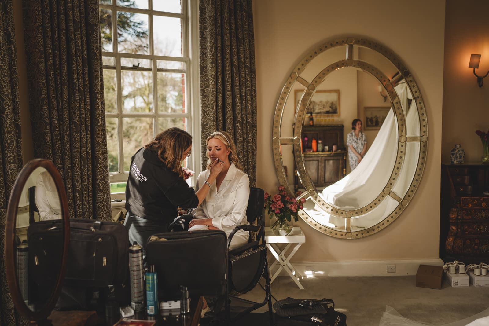 A woman is having her makeup done in front of a large mirror at Iscoyd Park. In the background, another person holds a white wedding dress, ready for her big day in Shropshire.