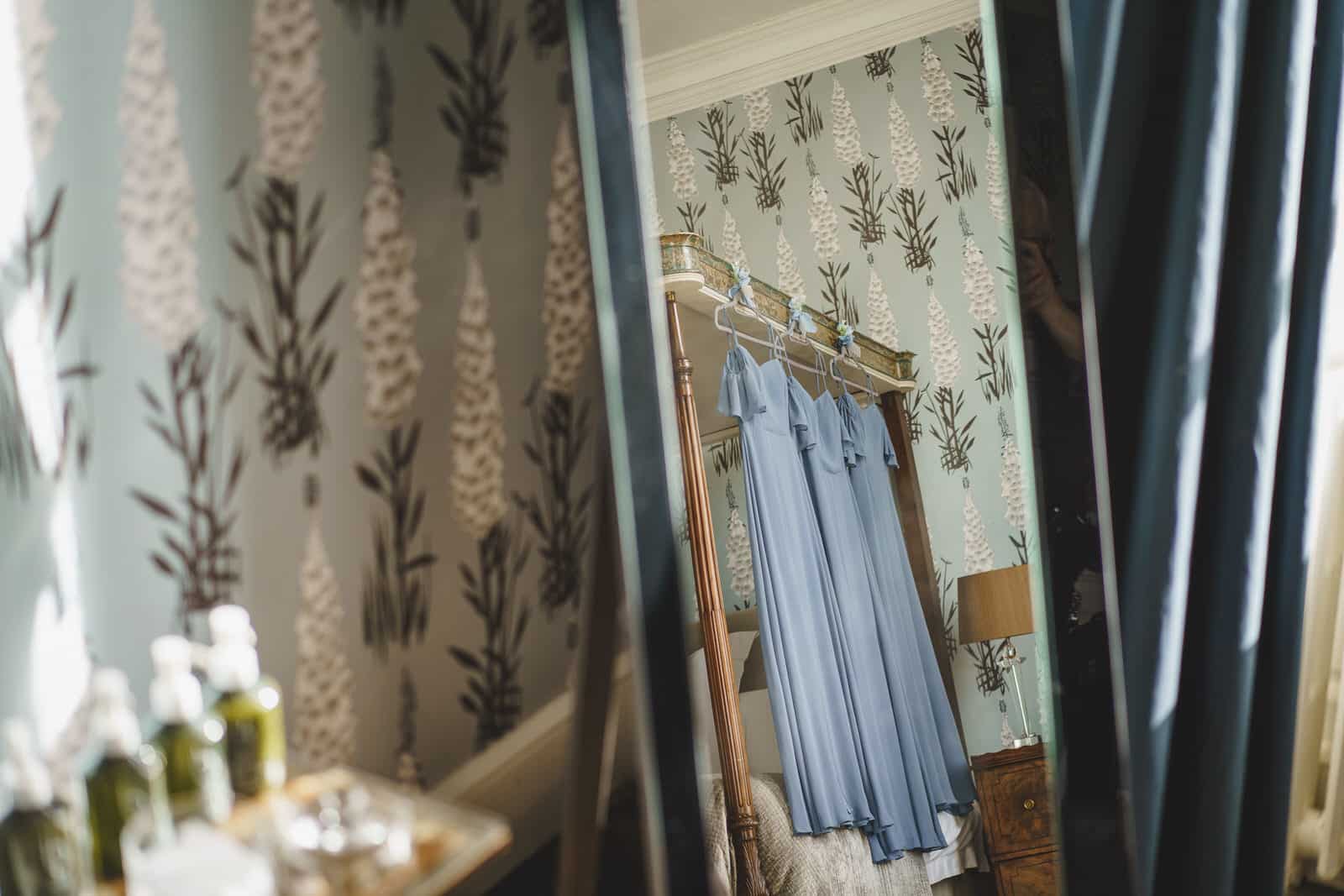 Room interior with floral wallpaper, a bedside table, and blue dresses hanging on a bedpost. Reflections are seen in mirrors or glass panels, capturing the enchanting ambiance of an evening at Iscoyd Park in Shropshire.