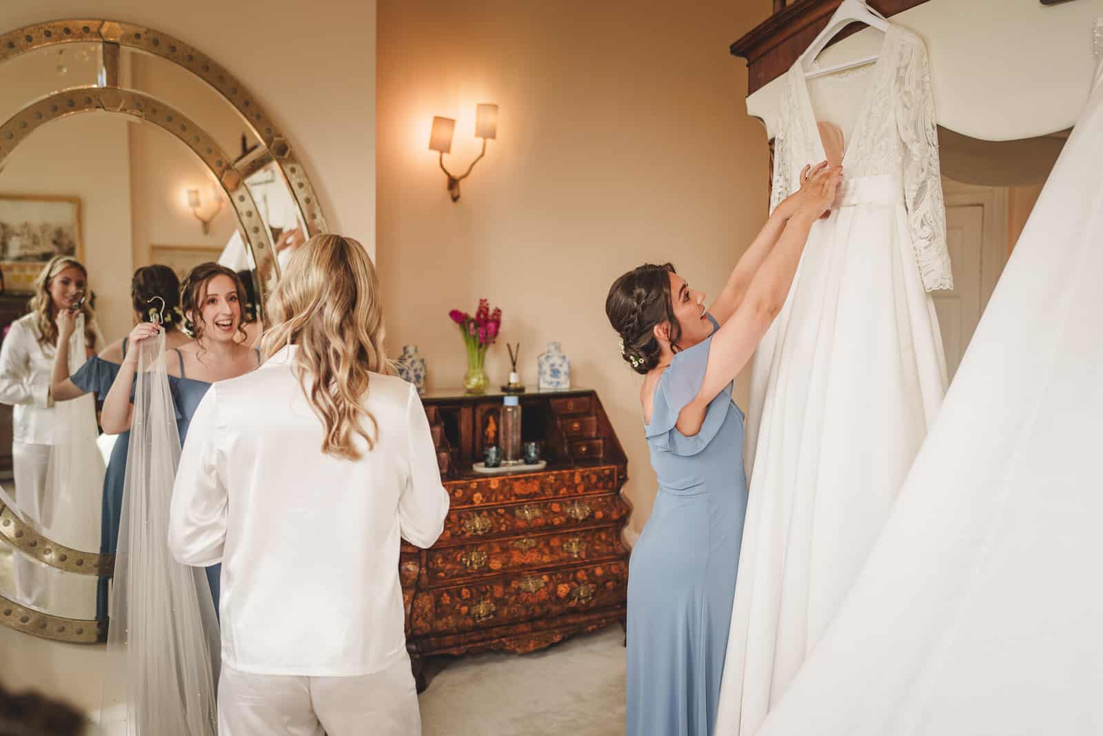 A woman in a blue dress holds up a white wedding gown while another woman in white stands in front of a mirror at Iscoyd Park. Two other women chat in the background of the warmly lit room, which features elegant wooden furniture, capturing a cozy Shropshire charm.